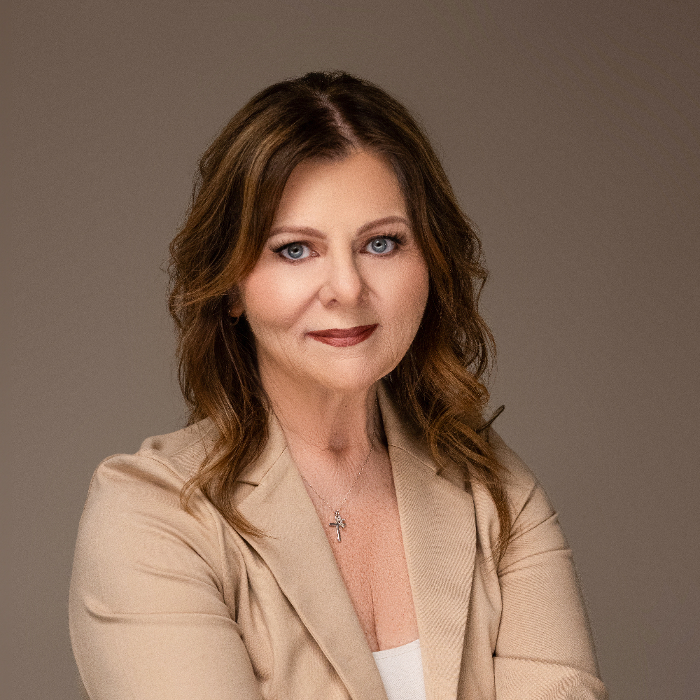 A woman with long hair wearing a beige blazer and a white top, looking directly at the camera.